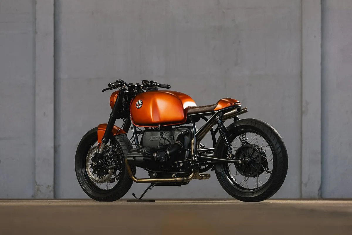 BMW R100/7 cafe racer naranja de Caffeine Custom vista de perfil junto a un muro de hormigón, con el motor bóxer a la vista, asiento monoplaza de cuero marrón y escape de acero inoxidable