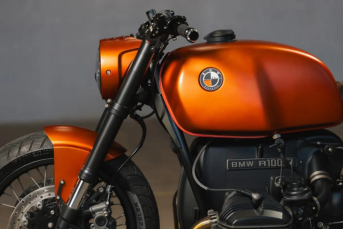 Detalle del lado izquierdo de la BMW R100/7 naranja: depósito original con la insignia redonda, guardabarros delantero a juego, horquilla negra y la placa «BMW R100/7» del motor