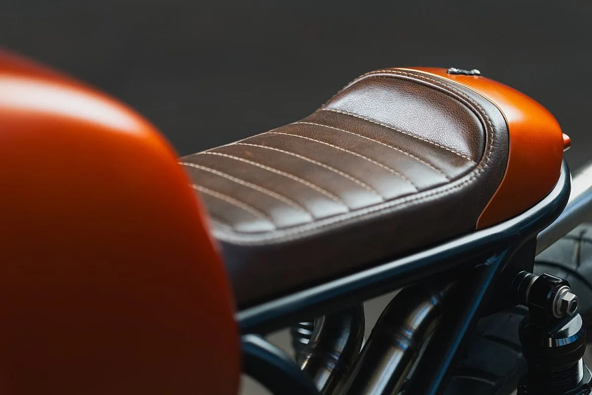 Asiento monoplaza de cuero marrón capitoné con costuras claras sobre base de aluminio, junto al depósito naranja de la BMW R100/7