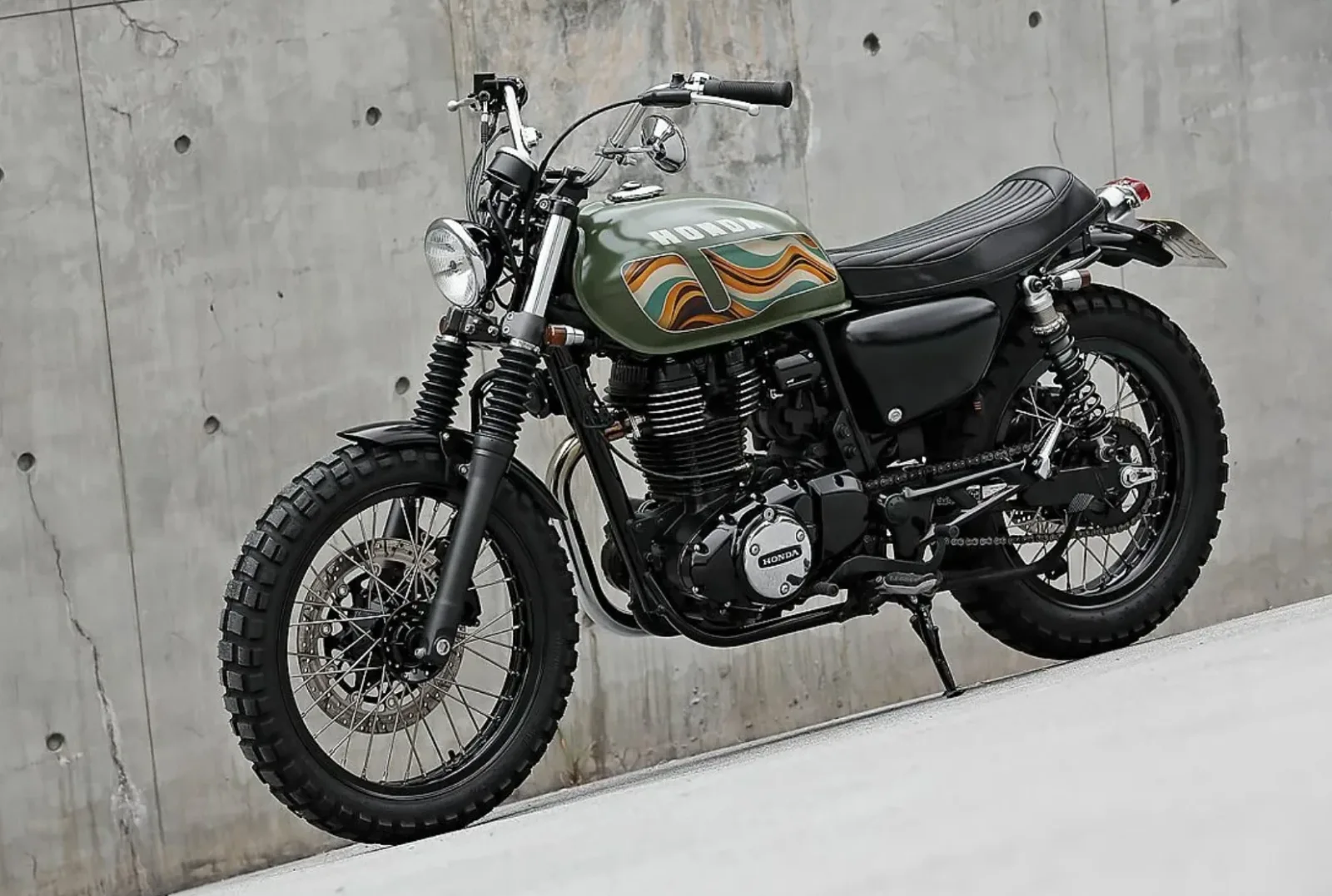 Honda CB350 scrambler en tres cuartos delanteros: faro redondo, horquilla con fuelles, guardabarros negros y neumáticos de tacos delante de un muro de hormigón