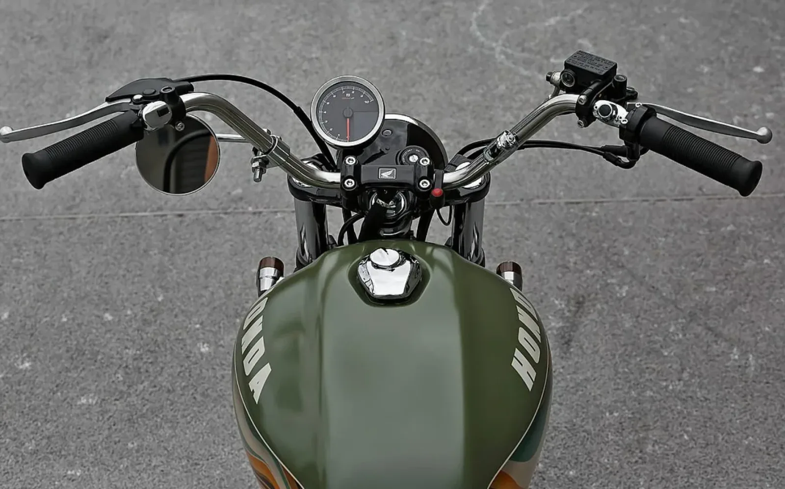 Vista desde arriba del manillar alto de la Honda CB350 scrambler, con el velocímetro redondo, un retrovisor circular y el depósito verde con el nombre Honda