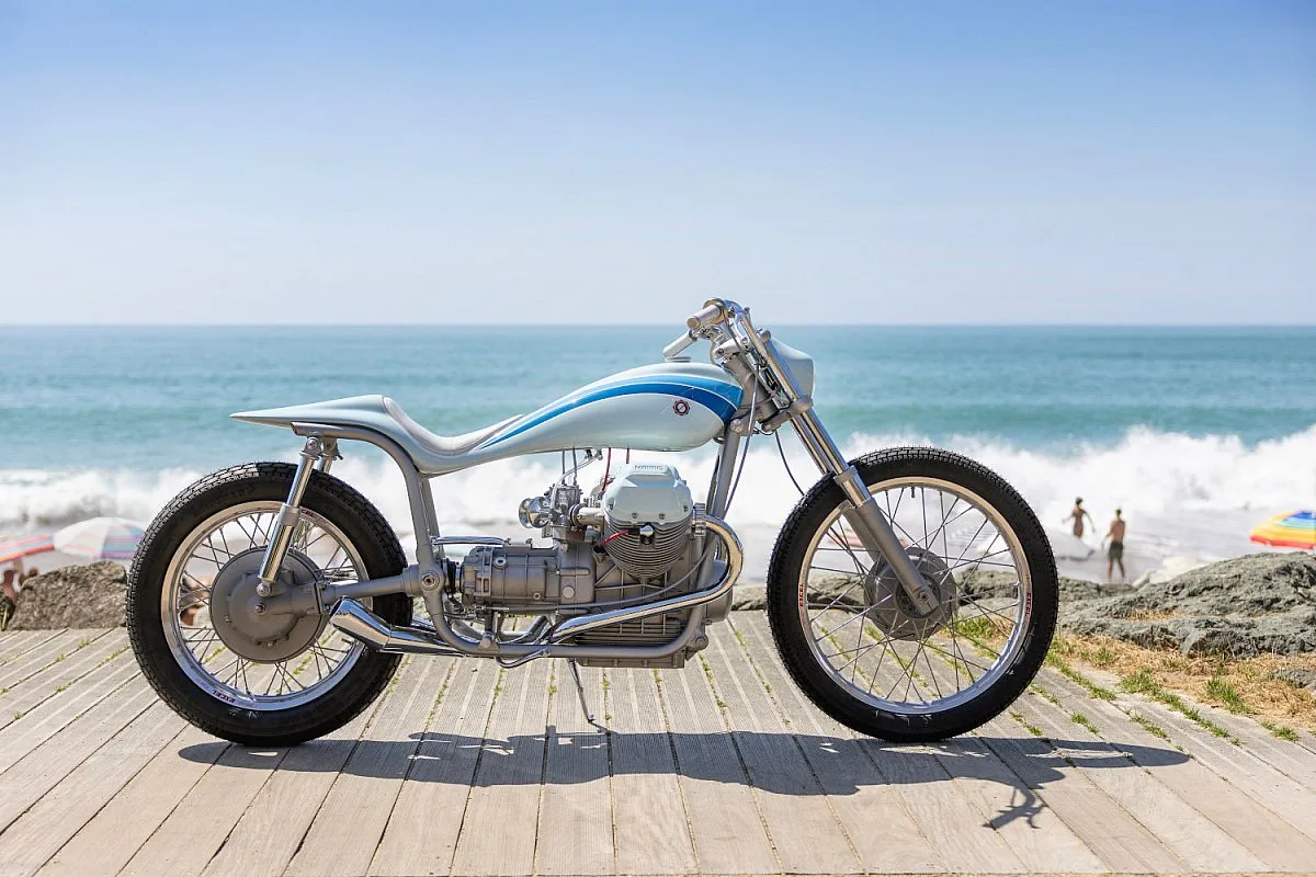 Moto Guzzi V7 «Bonanza» de Kaffeemaschine vista de perfil en un paseo marítimo: depósito y colín plateados de una pieza con franjas azules, motor bicilíndrico en V a la vista y ruedas de radios