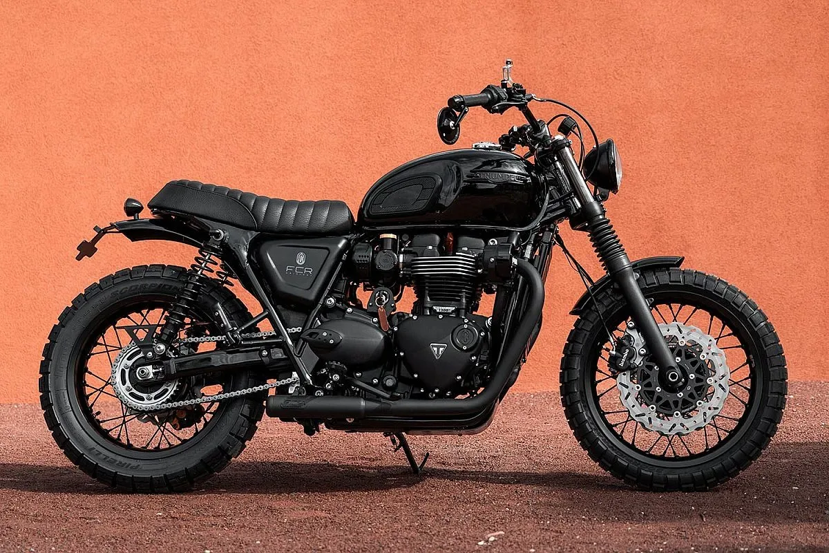 Triumph Bonneville T120 scrambler de FCR Original, vista de perfil: moto toda en negro con asiento acolchado, neumáticos de uso mixto y llantas de radios, frente a un muro naranja